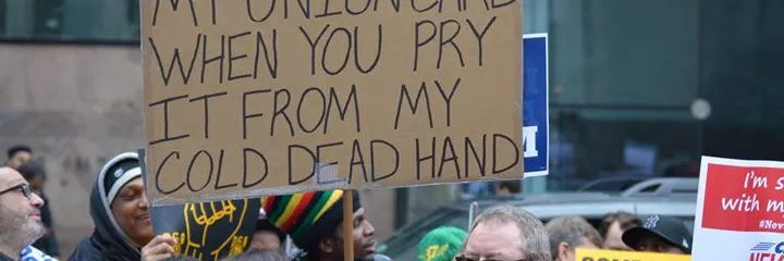 man holding sign in a crowd that says "i'll give up my union card when you pry it from my cold dead hand"